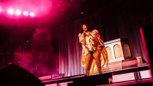 Lizzo performs in a shiny gold robe on stage at the Yuengling Center under pink colored lights
