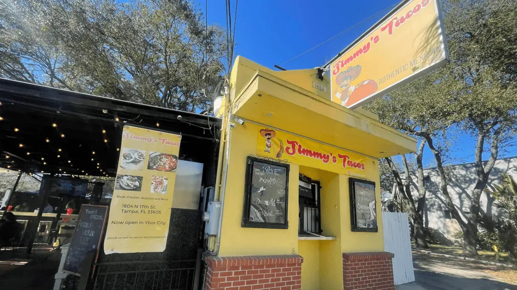 Exterior of a yellow restaurant. A sign reads tacos over the order window. Some outdoor patio seating is visible.