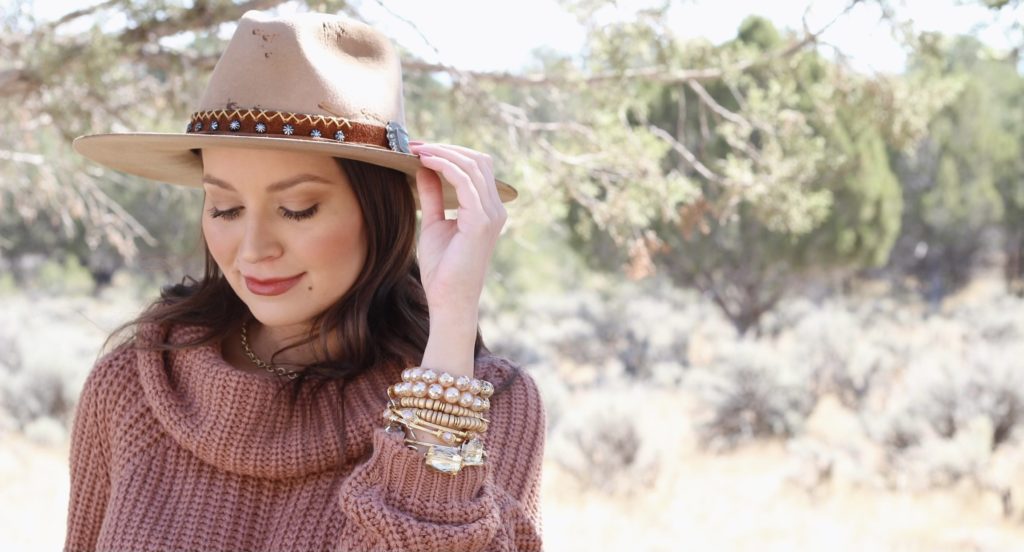 a brunette woman wearing a sweater models a cowboy style hat and stack of various bracelets