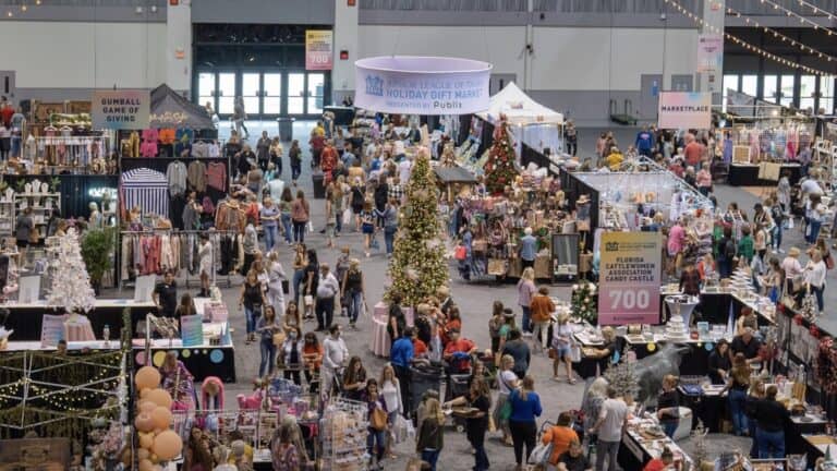 A bustling indoor holiday gift market with decorated Christmas trees and numerous vendor booths, attracting a crowd of shoppers.