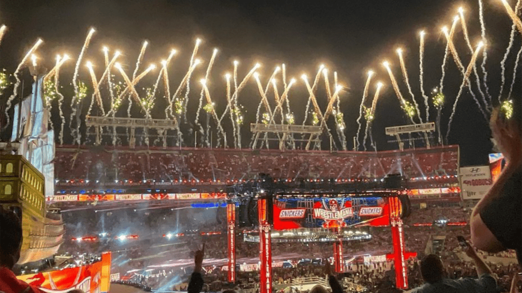 an open air stadium with fireworks going off in the background at night with a giant screen and pirate ship set in the corner