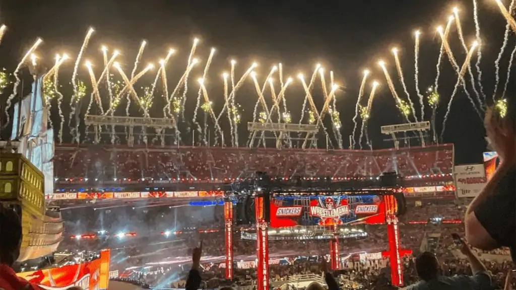 an open air stadium with fireworks going off in the background at night with a giant screen and pirate ship set in the corner