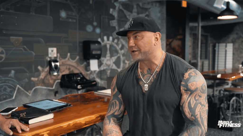 A man with tattoo sleeves since inside a tattoo studio with large wooden tables next to him.