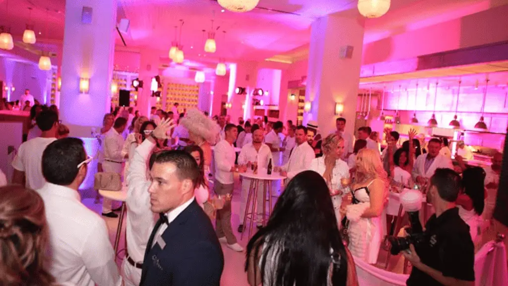 Interior of a large bar area bathed in pink light. Attendees to a party are dressed in all white.