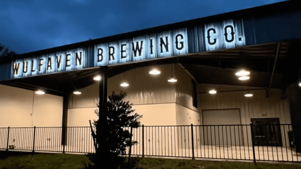 Exterior of a brewery. Open air sitting is shown. A blue neon sign spells out the name of the brewery