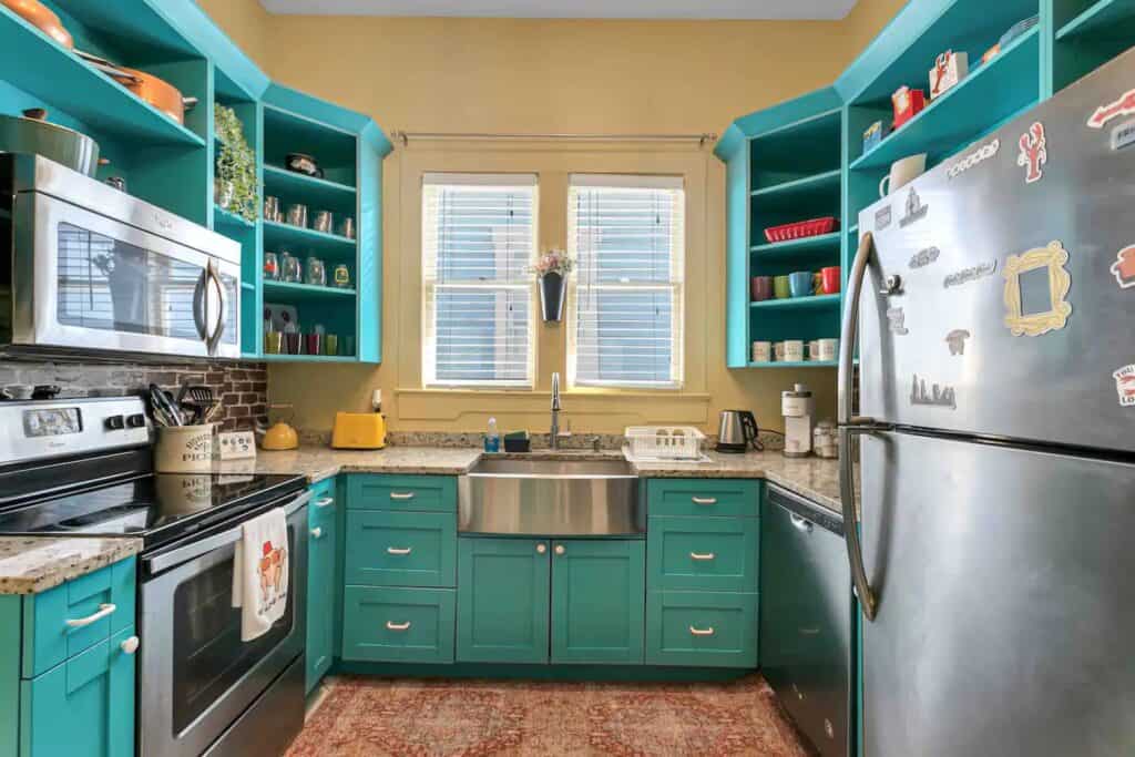 a kitchen with turquoise drawers and cabinets. A stainless steel refrigerator , and colorful mugs. arranged on shelves.
