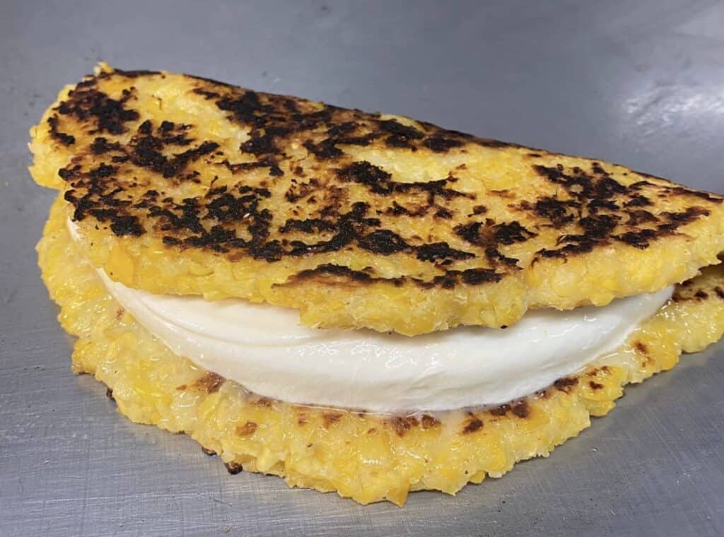 A fried corn patty folded over a block of cheese on a griddle. 