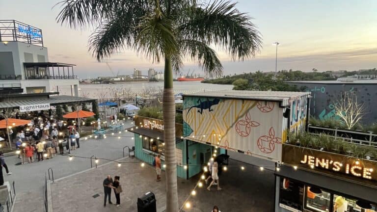 An outdoor waterfront food hall wit shipping containers acting as vendor stalls. String lights connect the shipping containers. A tall palm tree is at the center of the photo.