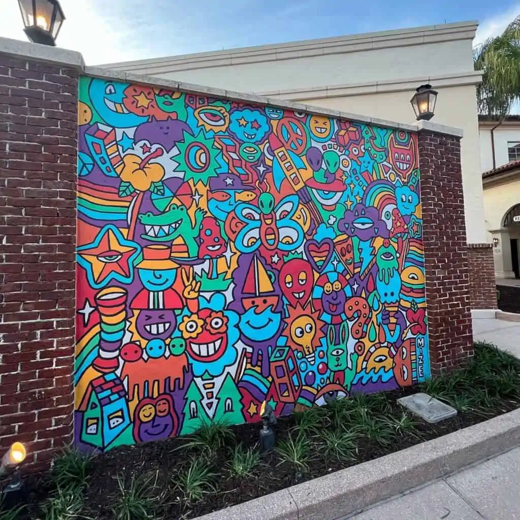 A colorful mural featuring animated plants, animals, sail boats and more. Green, purple, red, orange, and blue are the prominent colors on the mural It's framed by two brick pillars with lamps on top. 