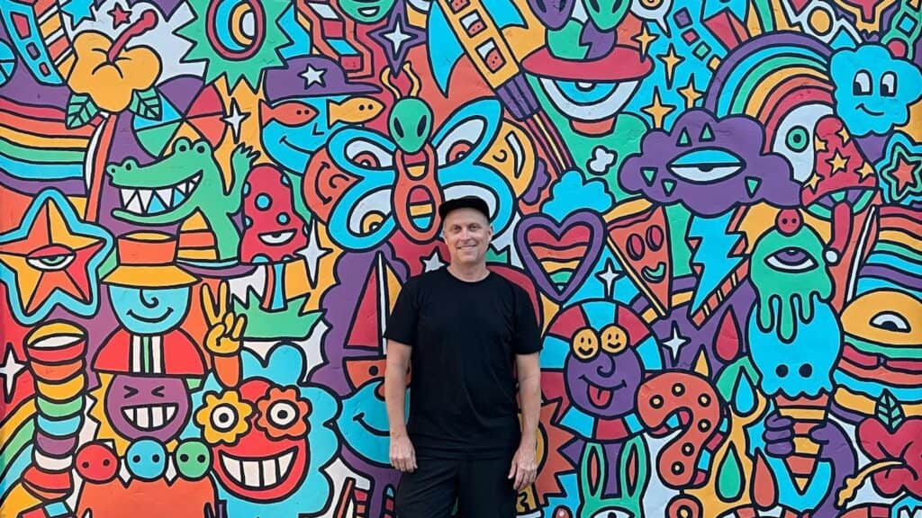 an artist in all black, wit ha black baseball cap on, stands in front of a rainbow colored mural with carton animals painted on it