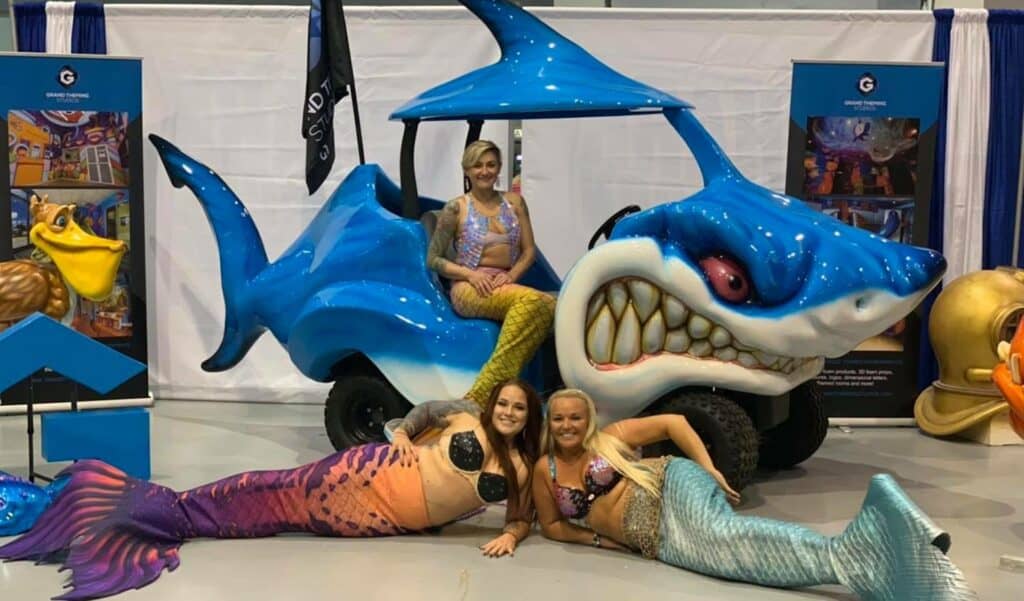 A large car with the body pf a shark is painted brilliant blue. Three mermaids sit in front of the vehicle. 