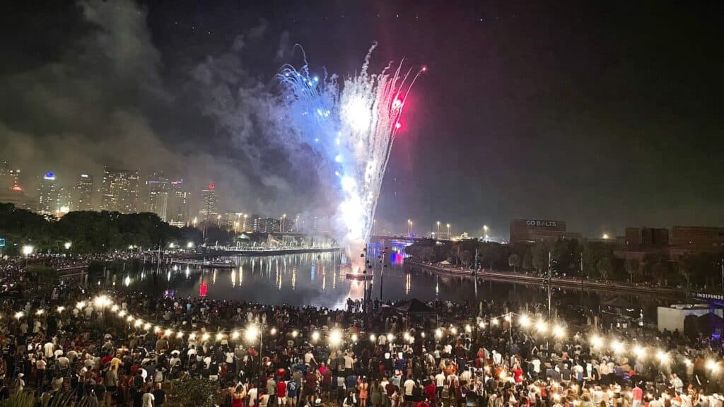 fireworks display the waterfront. String lights hang over a capacity crowd on a long riverwalk