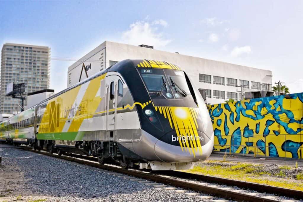 Photo of a train at a stop with a concrete wall. The wall is painted blue and yellow. The trains sun streaks painted on the side. 