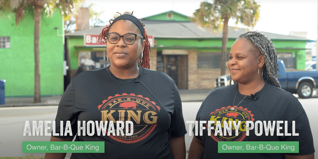 Two women in t-shirts that read "Bar-B-Que King" stand in front of a small restaurant. 