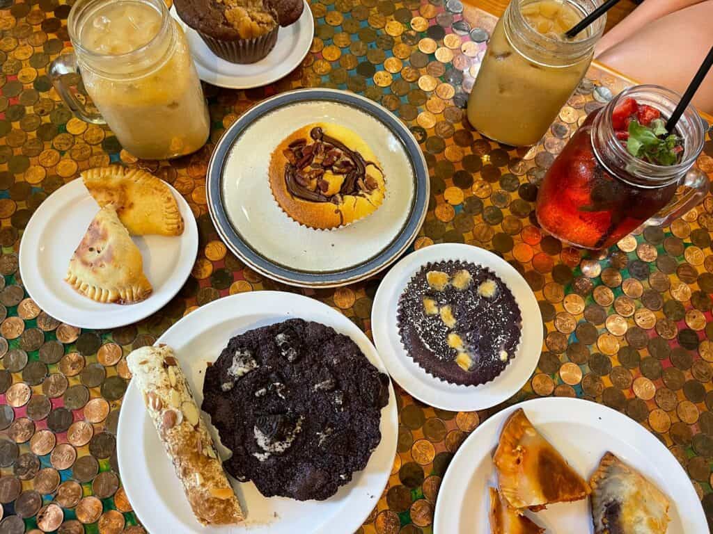 A table with an array of pastries set up on it. The table is fixed with hundreds of pennies waxed and glued to the top. A red spritz drink is set next to a latte, both are in mason jars. 