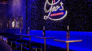 interior of a champagne bar. An ivy wall features a neon sign. Champagne bottles are lined up on tables.
