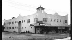 Black and white photo of a three story building. An old car is driving off the distance.