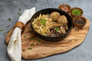 a bowl of beef meatballs, mushrooms, and pasta sits on a wooden block
