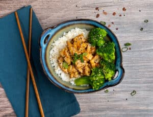 An aerial of a bowl of rice, broccoli, and sesame chicken in a bowl next two a napkin and chopsticks