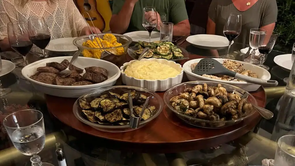 Variety of dishes served for dinner on a round table including steak, mashed potatoes, eggplants, mushrooms, corn on the cob, fish, zuchini