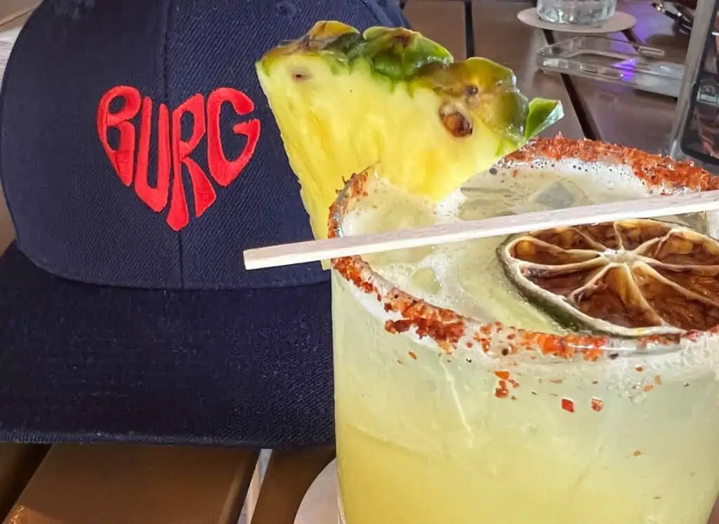 A glass of margarita with pineapple garnish next to a cap with text Burg in the shape of a heart