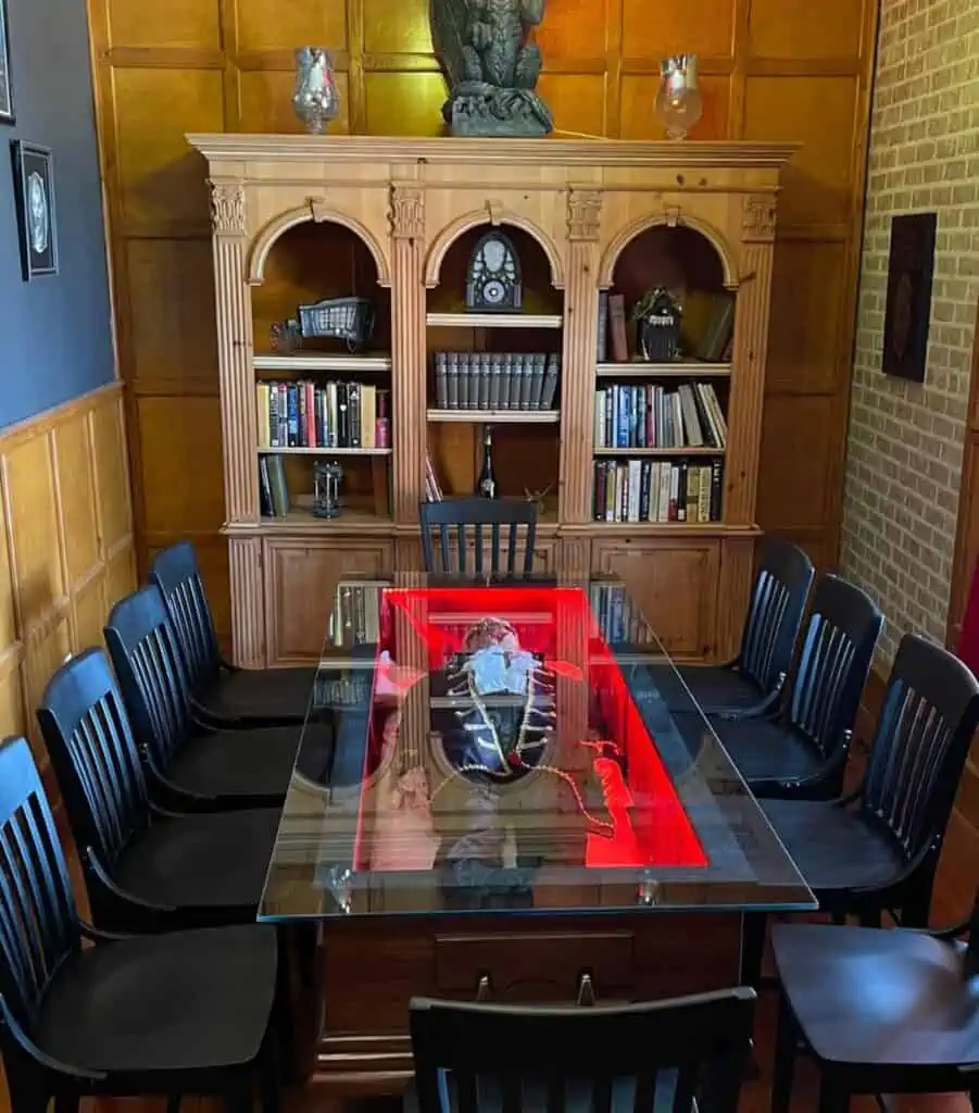Inside the SpookEasy meeting room. A glass plate is set over an old coffin with a figure inside dressed in Victorian fashion. Black leather seats surround the table and a bookcase is visible in the background. 