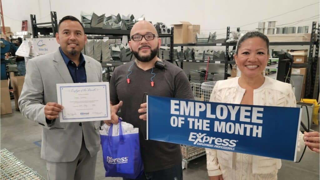 Three people in a warehouse. A woman in a white jacket holds a sign that read employee of the month. A man in the center with glasses on and a grey shirt holds a small blue gift bag.