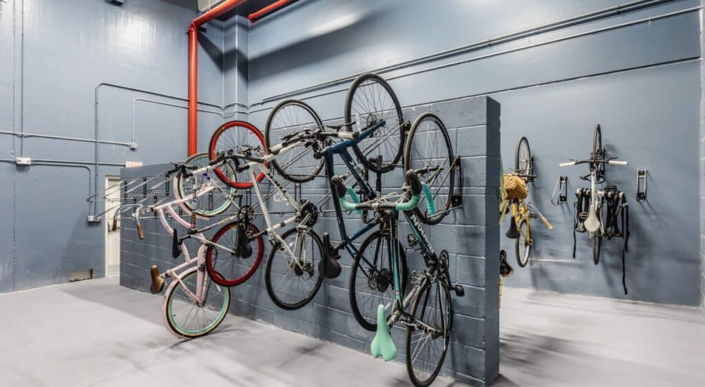 A large bike room. Roughly 8 bikes are set vertically along a grey brick wall. 