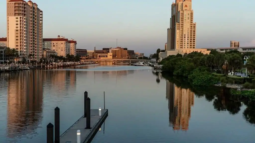 A view of a city skyline separated by a long river. A park is visible on the right side with a long brick walkway stretching from it.