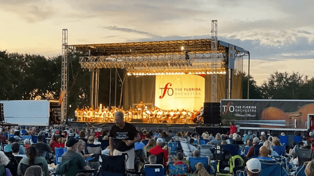 A stage in a park with an orchestra set up to perform. Attendees are seating in picnic and lounge chairs in the grass