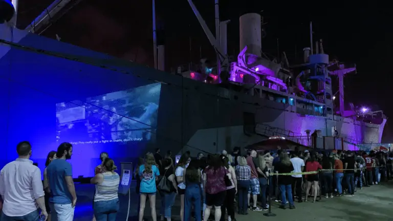 a group of people line up outside a historic ship lit up with blue LED lights