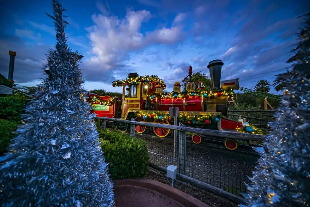A train covered in ornaments and ivy rides through an area replete with Christmas trees. 