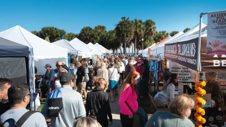 A crowded outdoor market features rows of vendor tents selling a variety of goods, with people browsing under a clear blue sky.