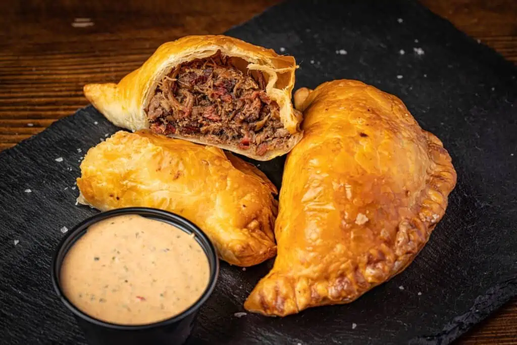 two empanadas on a plate with dipping sauce in a small cup. One empanada is open revealing brisket on the inside. 