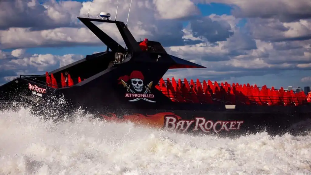 A speed boat with a skull and bones design on the side crashes through waves. The vessel is black with bright red seats