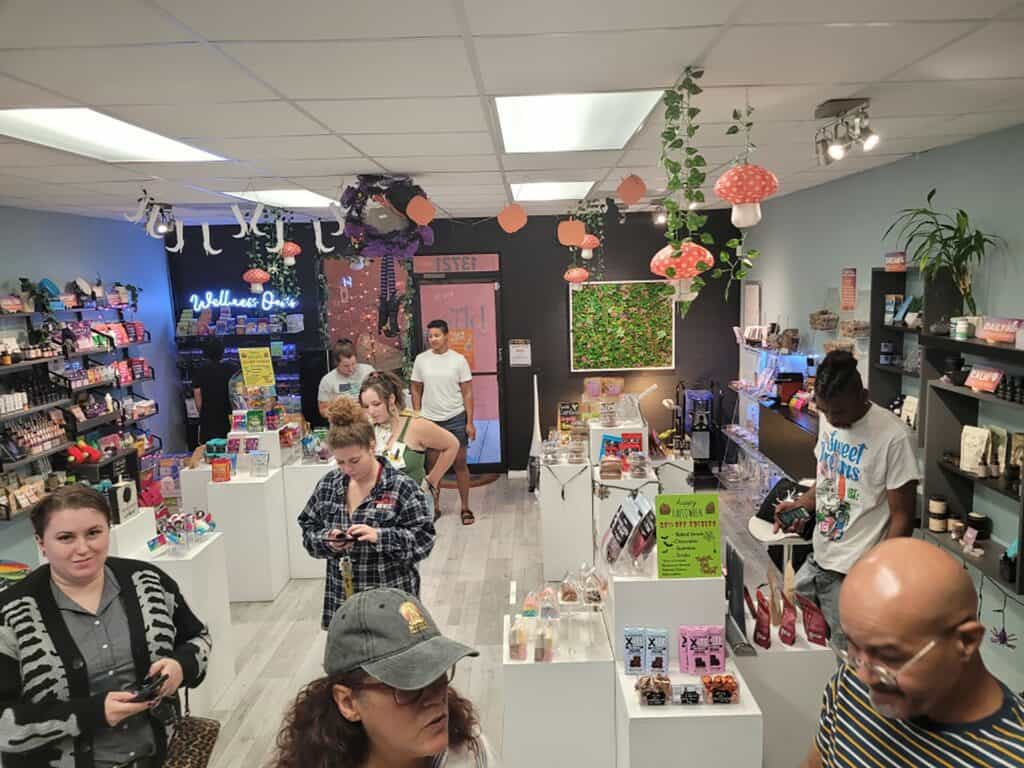 Customers gather inside a boutique wellness shop. Ivy and mushroom decor hangs overhead. Products are displayed on a array of white tables. 