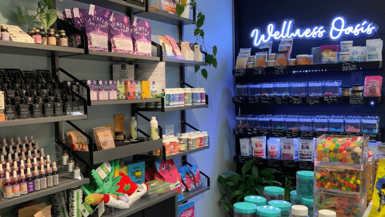 Interior of a CBD store. A neon sign that says wellness glows blue in the background. Gummy candies in tall glass jars are displayed on a wooden table. Three tiers of different snacks are arranged on a wire rack to the left of the photo.