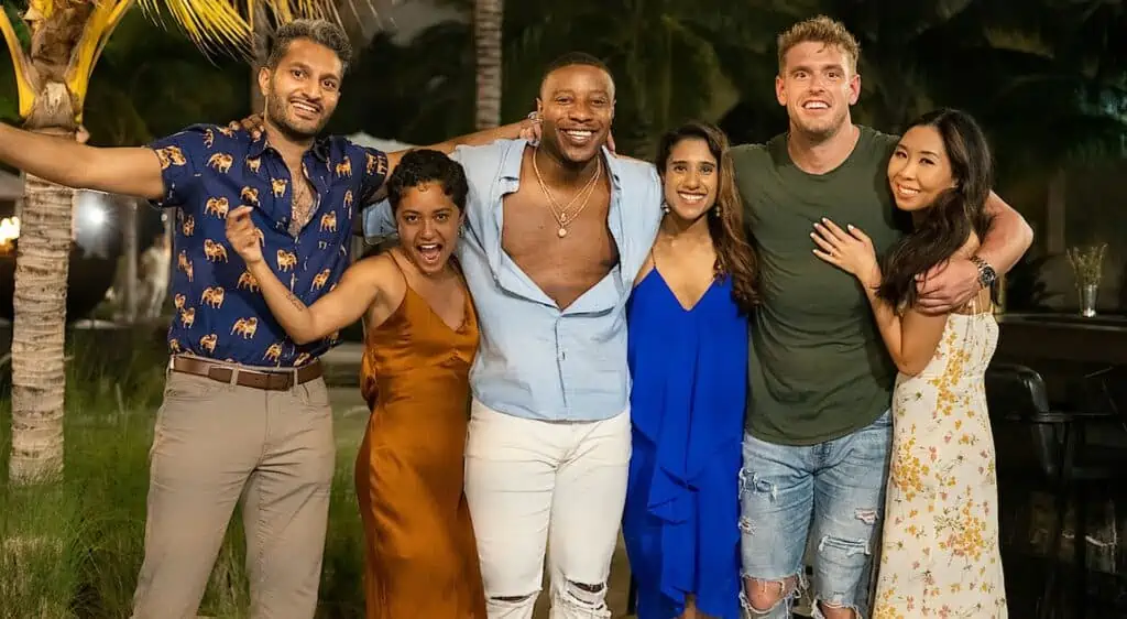 A group of people dressed in resort fashion wrap their arms around eachother. There are six people, three men and three women, in the photo.