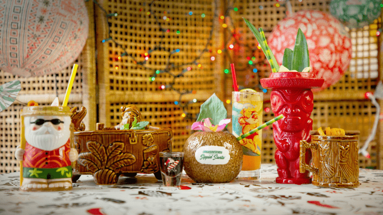 An array of craft cocktails displayed in Christmas-themed cups. Colorful Christmas lights hang in the background.