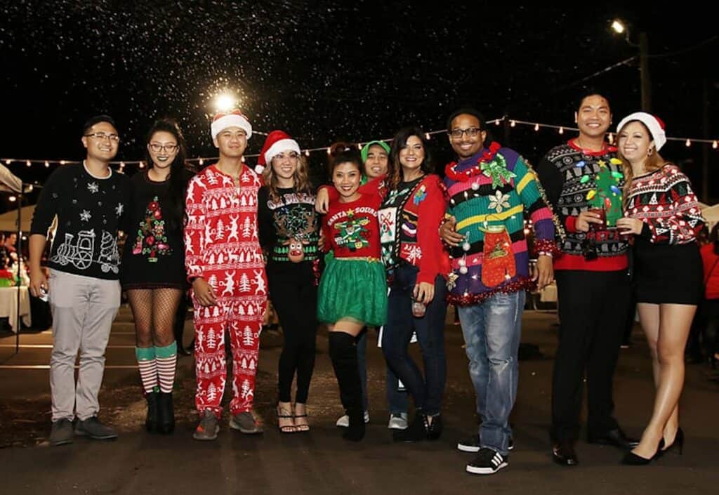 A group of people are gathered in a parking lot in a array of vintage ugly holiday sweaters. String lights hang overhead. There are nine people total in the photo. 