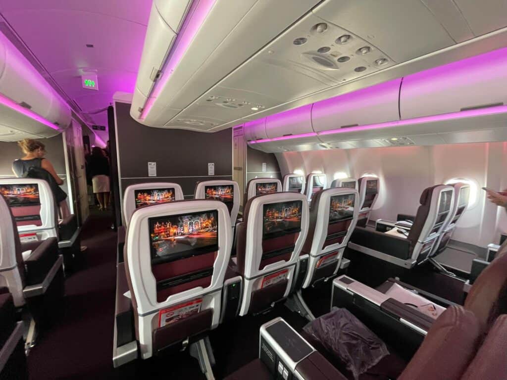 on a plane with multiple rows of luxurious seats and screens on the back of each chair. The airplane cabin is lit in purple light