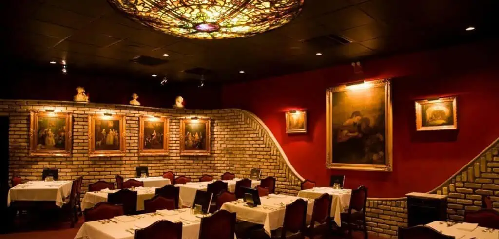 Inside a dining room with classic paintings hung on the wall. A red paint job is set next to brick walls. Tables are draped in white linens. 
