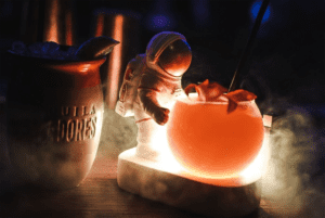 A cocktail glass that is smoking. an astronaut figured is placed next to the cup.