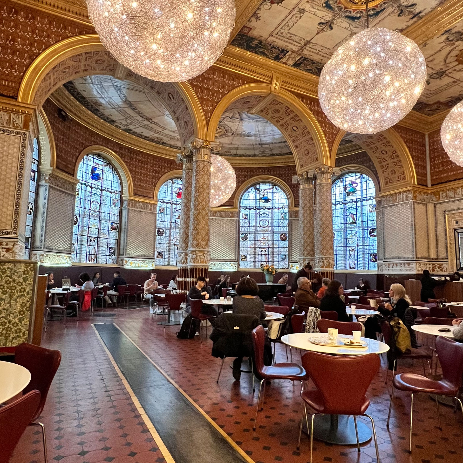 View of the cafe inside the Victoria and Albert Museum in London with people eating on cafe tables surrounded by ornate chandeliers and historic architecture