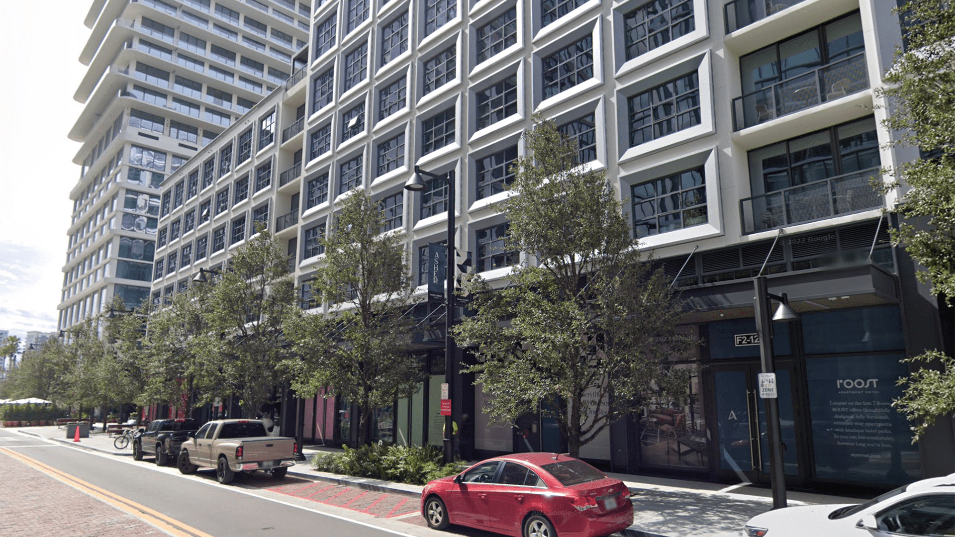 A street scape. Cars are parked on the side of the road. A brick patterned sidewalk runs down the center of the road. Trees provide shade on the sidewalk.