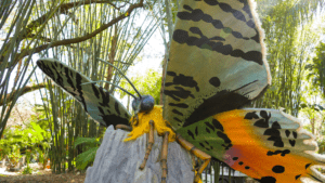 Giant butterfly robot with green wings and an orange body rests on a boulder in the woods
