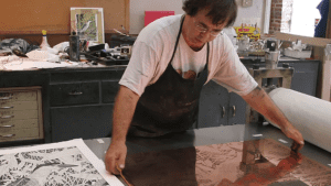 An artist in a white shirt and apron stands over a large copper plate.