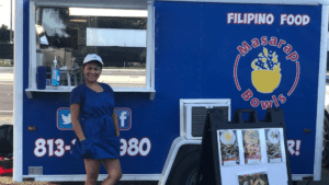 A person standing in front of a blue food truck with a yellow bowl painted on the side of the truck.