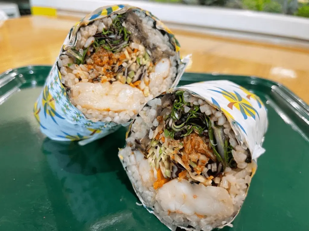 a sushi burrito in a while and yellow wrap loaded with rice, cabbage, kimchi and tofu on a green tray. 