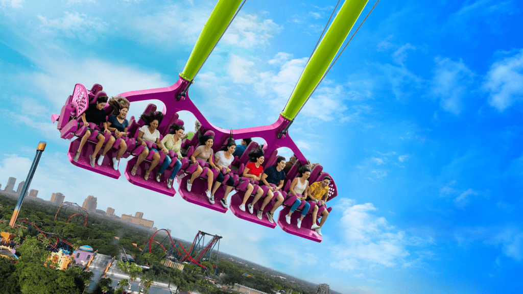 riders are strapped in on a large purple swing. It soars over an amusement park.